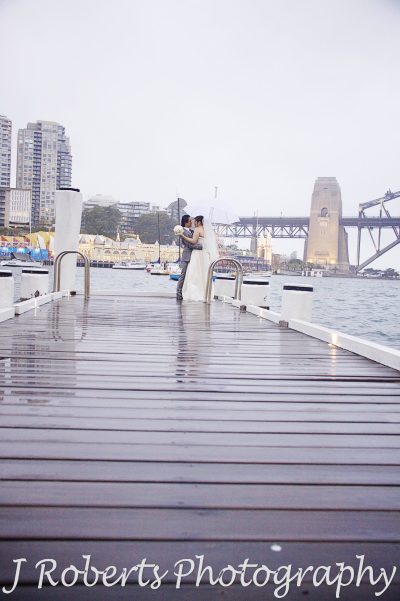 Couple embracing on pier overlooking Sydney harbour in the rain - wedding photography sydney